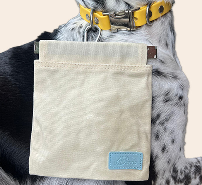 Beige canvas dog treat bag with blue Oh Vienna logo, featuring a squeeze pouch closure and a carabiner clip to attach to a dog’s collar D-ring